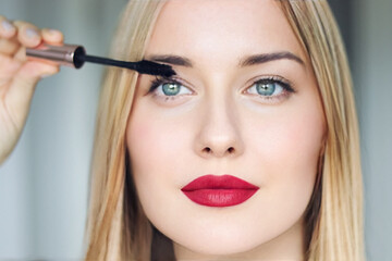 Woman applying mascara with confidence, defined brows and red lipstick. Blonde beauty focused on...
