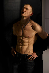 Fototapeta premium Slim muscular male model on black background. Shirtless fitness guy in black sweatpants posing in studio. Man with six pack abs in sunset light. Brutal photo