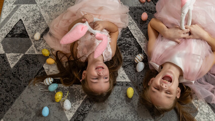 Cute laughing little girls with bunny ears are lying on a floor with colorful eggs in the Easter holiday.