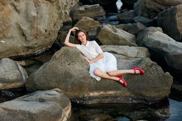 serene woman in white dress sits on rock in water, red shoes peeking out