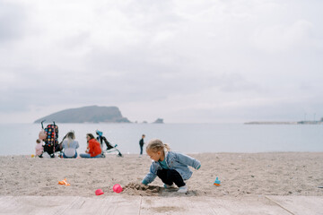 Little girl digs a hole in the sand while squatting against the background of mothers with children...
