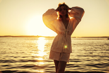 Young beautiful hipster woman in white dress posing  on sea background. Trendy model over sunset....