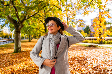 Happy middle aged woman in black brimmed hat standing in city park on autumn day. Front view	
