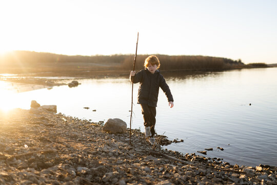 Boy walking along waters edge with long stick on sunny day