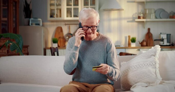 Medium portrait of aged male person with mobile phone and credit card at home. Retiree calling to bank, telephone scammers attacking lonely old people, security of financial data and bank savings