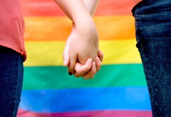 A close-up image of lesbian couple holding hands with a vibrant rainbow pride flag in the background, representing LGBTQ+ pride and inclusivity