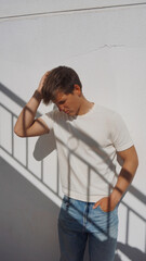 Young man touching his hair leaning on white wall outdoor, shadow of stairs on him