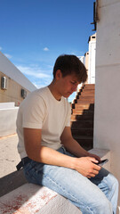 Teenage guy holding mobile in hands and sitting on wall on street