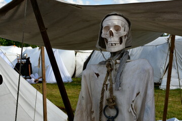 A close up on a replica of a skeleton made out of plastic, wearing peasant clothes and a modest hat, standing next to a set of white cloth tents during a medieval festival organized in Poland