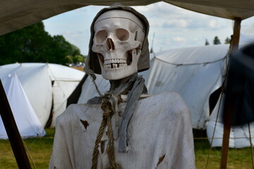 A close up on a replica of a skeleton made out of plastic, wearing peasant clothes and a modest hat, standing next to a set of white cloth tents during a medieval festival organized in Poland