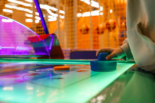 Child playing air hockey on a glowing table in an amusement center, close-up of hand holding paddle in neon light