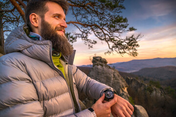 Bearded man in a puffer jacket stands on a mountain at sunset, checking time on Casio watch with a...