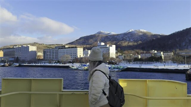 A person on a boat looks towards the town of Toyako Onsen on the shore of Lake Toya, with snow-capped mountains in the background under a blue sky