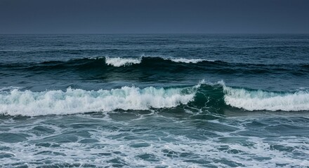 Fototapeta premium Blue ocean waves crash on the rocky coast, breaking with foamy surf near the sandy beach under a cloudy sky