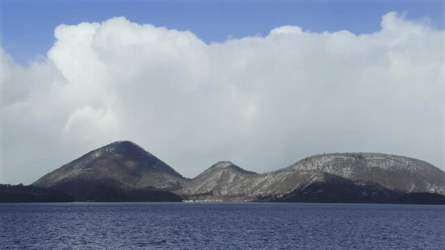 Snow-capped mountains rise from the tranquil waters of Lake Toya under a partly cloudy blue sky in Hokkaido, Japan