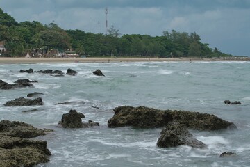 A beach with people playing in the water on a cloudy day, with rocks in the foreground