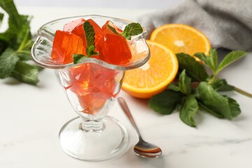 Delicious jelly cubes in dessert bowl, mint, spoon and orange on white marble table, closeup