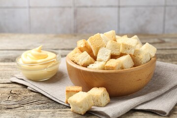 Tasty crispy croutons in bowl and sauce on wooden table against grey tiled wall, closeup
