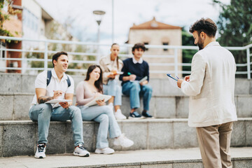 A group of students sit on outdoor steps, engaged in collaborative work with their professor. They...