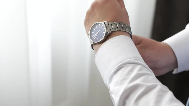 A man puts on a watch with a metal bracelet on his hand against the background of a window in a room