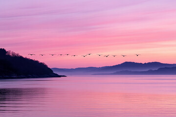 Birds flying in perfect formation over a calm lake at dusk surrounded by gentle hills and colorful skies