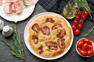 Delicious focaccia bread with bacon, rosemary, oil and tomatoes on black table, flat lay