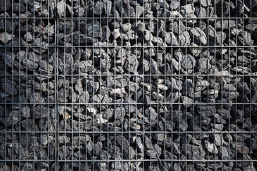 A tightly packed gabion wall consisting of rough gray rocks held together in a structured metal...
