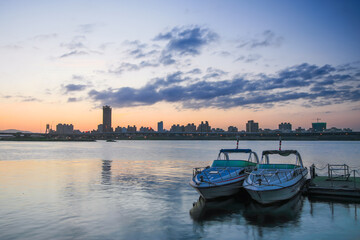 As the sun dips behind Taipei's skyline, Dadaocheng Wharf turns into a peaceful haven. Two boats rest quietly on the shimmering water, inviting serene moments under a dusky sky.