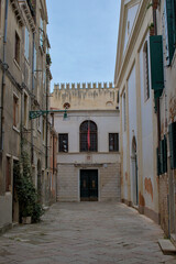 Fototapeta premium A quiet Venetian alleyway stretches into the distance, revealing a building with a red flag hanging above its dark double doors. Weathered walls hold stories of centuries past.