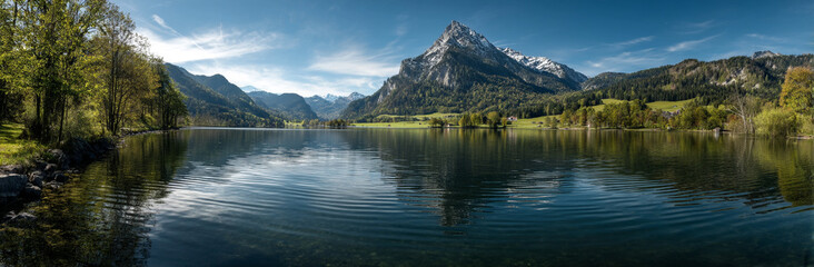 Panoramic photo of Lake. The lake is clear and calm, reflecting the surrounding mountains and trees in its surface. In front of it stands Mount Hood with snow-capped peaks against a blue sky backgroun