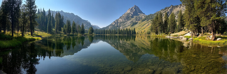 Fototapeta premium Panoramic photo of Lake. The lake is clear and calm, reflecting the surrounding mountains and trees in its surface. In front of it stands Mount Hood with snow-capped peaks against a blue sky backgroun