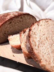 Freshly baked round bread cut into slices on a wooden board with a knife on a white fabric background. Close up.