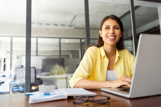 Young woman employee using pc for business work. Smiling latin female studying student, coder programmer working at laptop computer sitting at desk in modern office, dreaming looking at copy space
