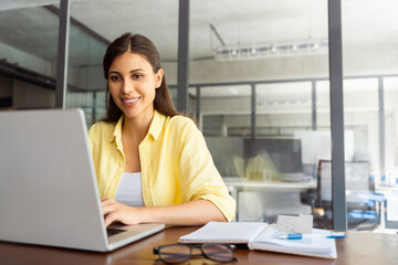 Smiling latin hispanic female marketing manager, professional it specialist working, browsing at laptop computer sitting at desk in modern office. Cheerful young woman employee using pc for business 
