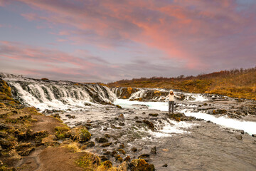 Long exposure photography of Brúarfoss Waterfall in Iceland beneath a dusk sky. Its turquoise stream cuts through volcanic terrain, framed by Highland mountains and autumn-colored shrubs
