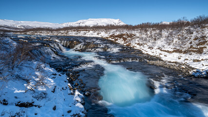 blauer Wasserfall Bruarfoss in winterlicher Landschaft bei strahlend blauem Himmel