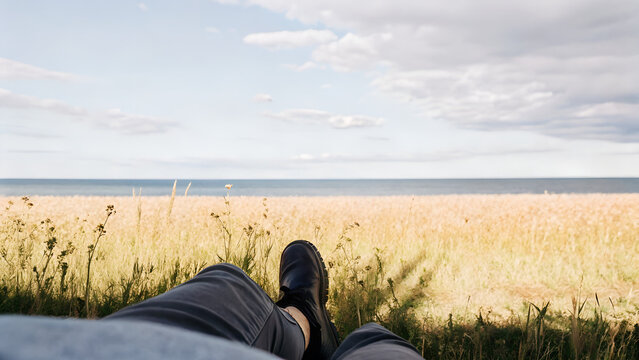 First-person view of relaxing near sea with legs in frame and horizon line over dry grass field ideal for travel blogs, relaxation-themed visuals, and summer social media templates