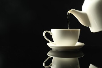 Pouring freshly brewed tea from teapot into cup at dark mirror table, closeup. Space for text