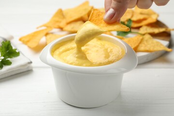 Woman dipping nacho chip into tasty cheese sauce at white wooden table, closeup