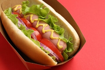 Tasty hot dog with tomato, onion, lettuce and mustard on red background, closeup
