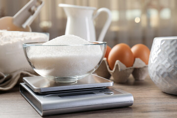 Digital kitchen scale with bowl of sugar among other products on wooden table, closeup