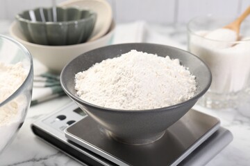Digital kitchen scale with bowl of flour and other products on white marble table, closeup