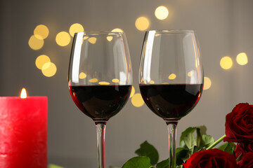 Glasses of red wine, burning candle and roses against blurred background, closeup. Romantic dinner
