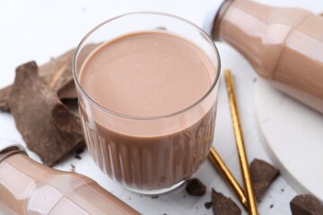 Tasty chocolate milk in glass, bottles and pieces of chocolate on white tiled table, closeup