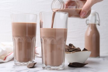 Woman pouring tasty chocolate milk into glass at white marble table, closeup