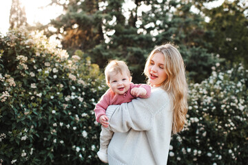 Smiling stylish woman holding baby girl 1 year old wearing knitted clothes over blooming bushes with flowers in meadow. Springtime. Motherhood. 