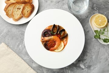 Delicious seafood soup in bowl, pieces of bread, water and lemon on gray textured table, flat lay
