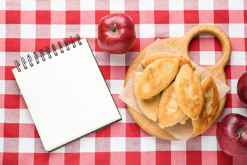 Blank recipe book, fried pyrizhky (stuffed pies) and apples on tablecloth, flat lay