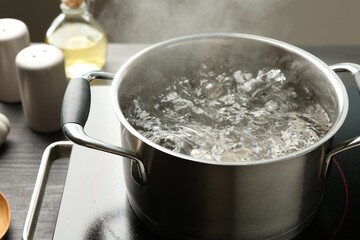 Cooking pot with boiling water and stove on wooden table against grey background, closeup