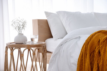 Bedside table with flowers in vase, glass of water and glasses near bed indoors